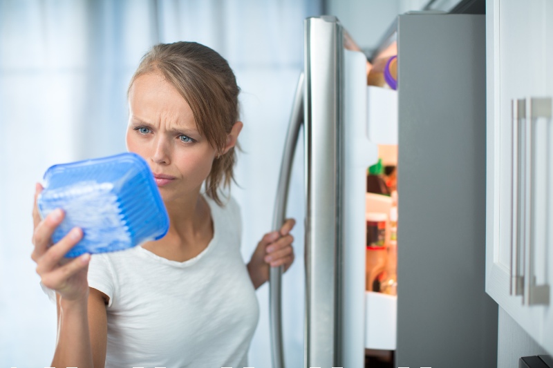young woman in her kitchen by the fridge, looking at the expiry date-Food Kit-SS