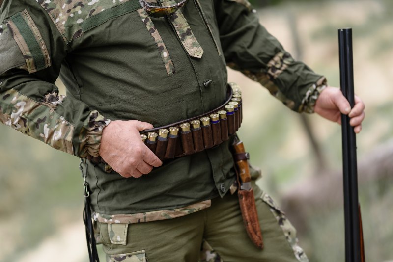 Hunter man stands with a belt of ammunition and a gun in his hands. A hunter with cartridges on his belt stands in the forest-battle belts