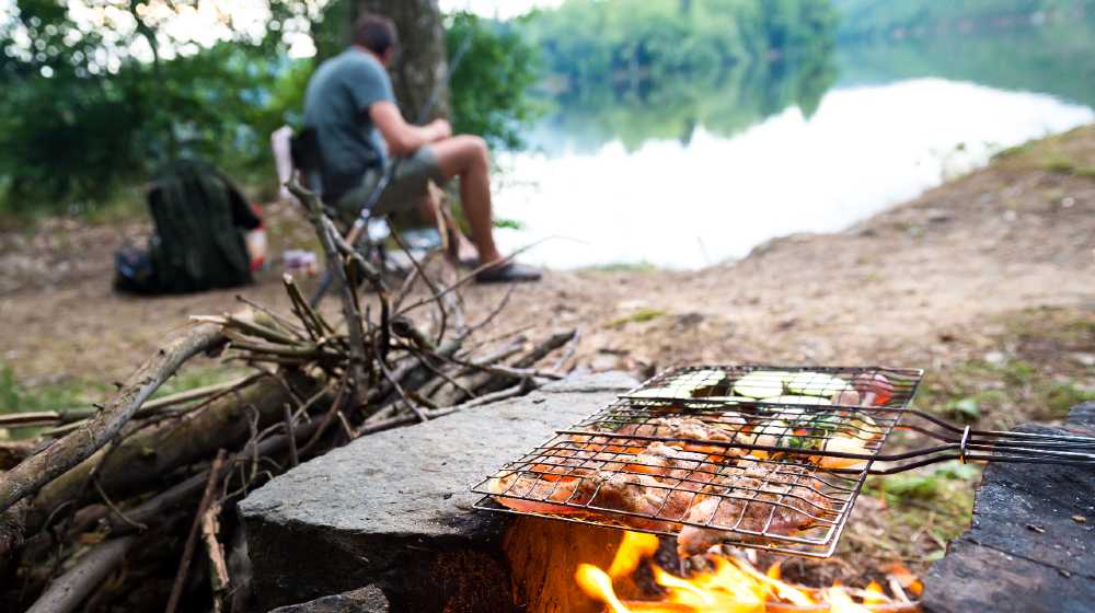 Fisherman preparing dinner on campfire, adventure lifestyle camping fishing vacation concept | Must-Try Campfire Fish Recipes For Your Next Outdoor Adventure | featured