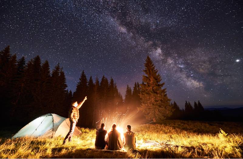 Male showing his friends Milky Way over camping. Guys are sitting by the campfire-terrain