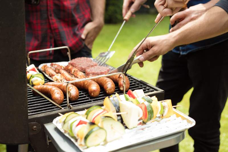 Outdoor shot of three friends enjoying barbecue party, grilling sausage, burgers and vegetable skewers-camping food hacks