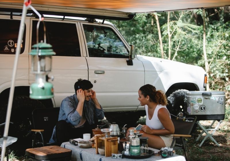 Positive multiethnic friends sitting at table near van | Tips to Help You Camp in the Rain