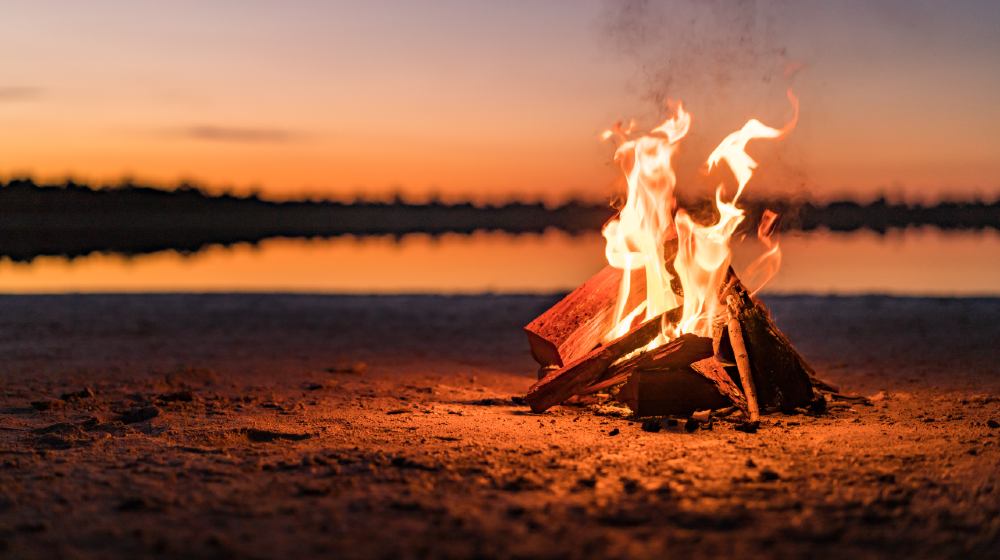 Small campfire with gentle flames beside a lake during a glowing sunset | The 5 Most Basic Types of Campfires And What They're For | featured