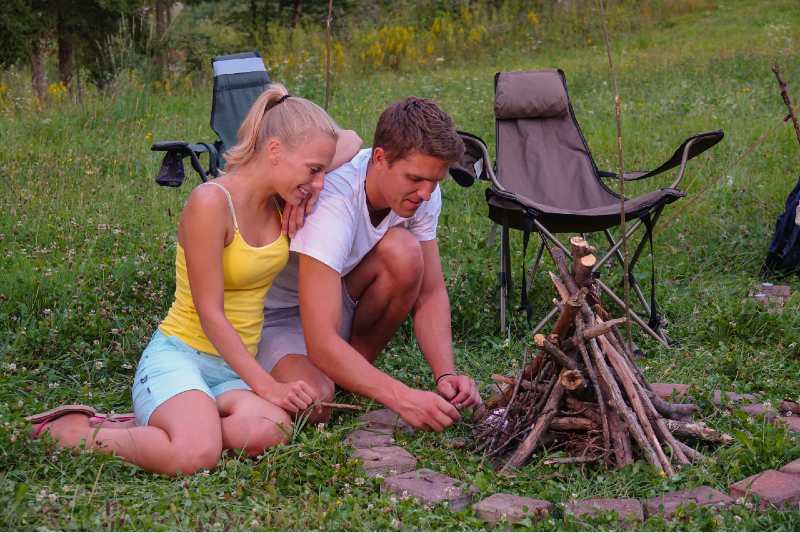 Smiling woman leans on her boyfriend who is lighting up the campfire by their tent-campfire