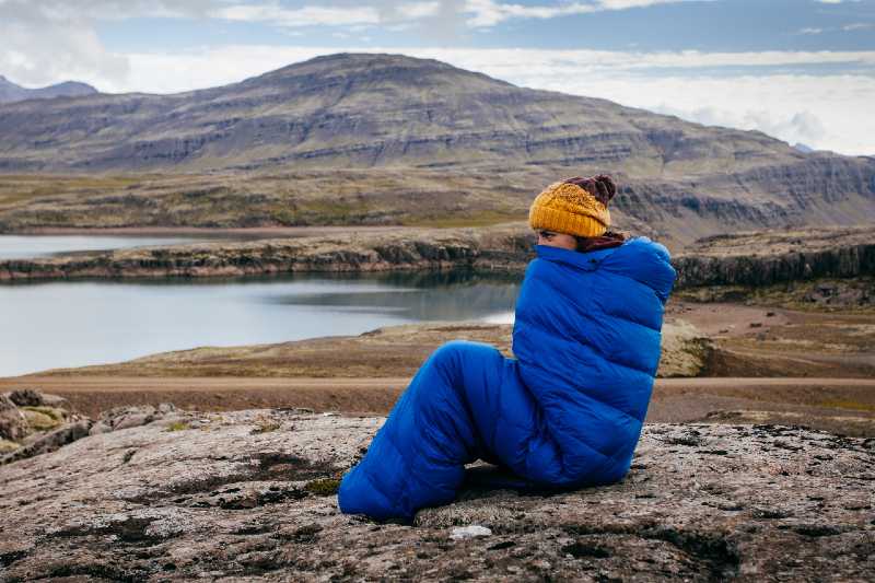 Young woman in warm blue sleeping bag , on the stones with northern mass-sleeping bag