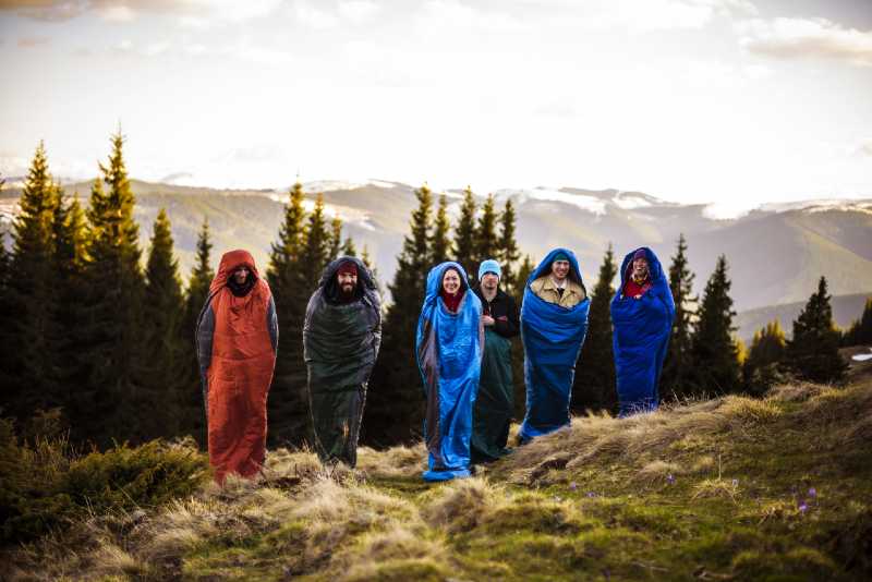 cheering group of hikers jumping in sleeping bags outdoors in mountains during the sunset-sleeping bag