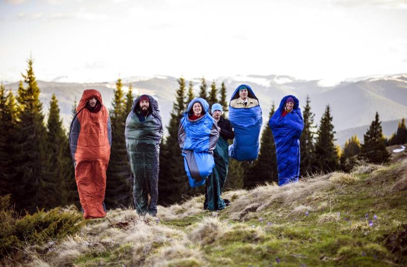 cheering group of hikers jumping in sleeping bags in outdoor mountains during sunset - find a campsite