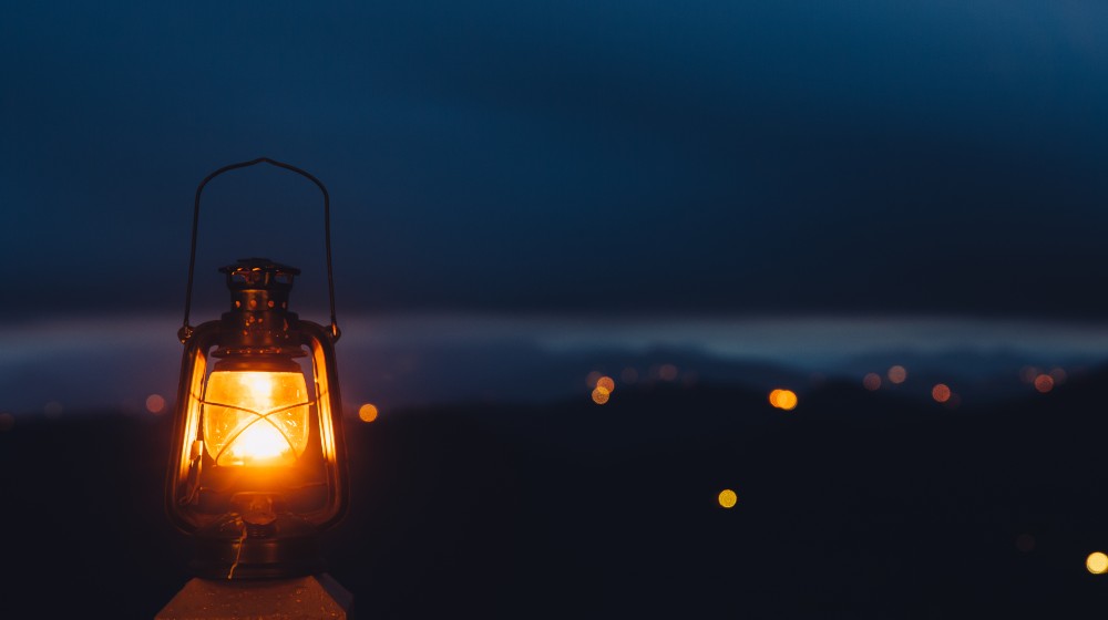 Blurred background image of a glowing lantern against dark night time mountains with lights | The Benefits of Having a Camping Lantern | featured