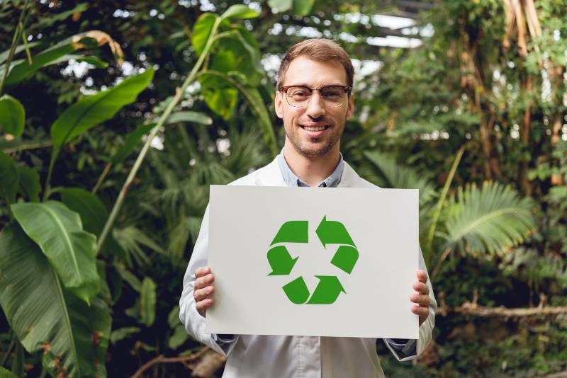 smiling handsome scientist in white coat and glasses holding card with green recycling sign in orangery | Environmental Policy
