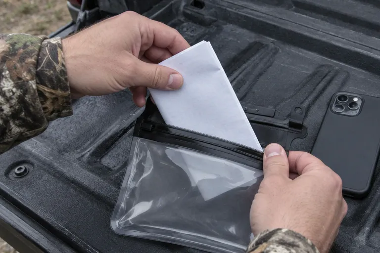 Hands sliding a hunting license into a clear waterproof pouch next to a phone on a truck tailgate